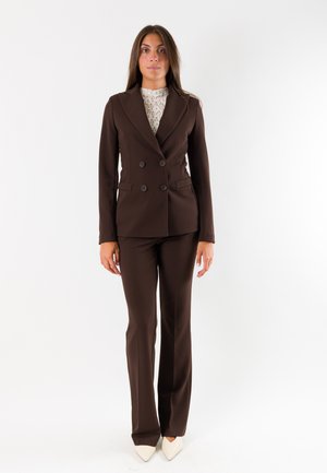 Donna in piedi che indossa un blazer doppiopetto aderente marrone scuro, pantaloni abbinati, top bianco in pizzo e scarpe bianche a punta su sfondo bianco.
