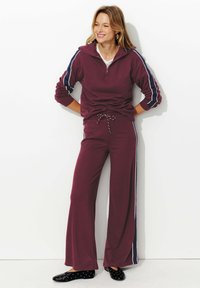 Survêtement bordeaux avec un col haut, fermeture éclair à l'avant et rayures latérales bleu marine. Pantalons à coupe ample avec cordon de serrage, associés à des ballerines noires à pois.