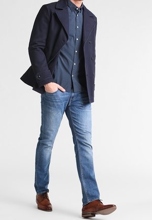 Man draagt een marineblauw dubbelbreasted jasje, een blauw hemd met patroon, lichtblauwe spijkerbroek en bruine leren nette schoenen, staand met één hand in de zak.