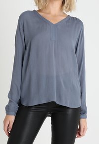 Blouse bleue à manches longues avec un décolleté en V, en tissu texturé, présente une coupe légèrement ample et un ourlet incurvé.