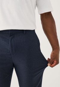 Personne à la peau foncée portant une chemise blanche à manches courtes étirant le tissu d’un pantalon bleu marine à fines rayures au niveau de la cuisse.