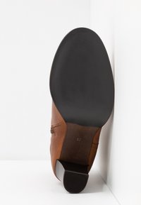Botte en cuir marron avec un bout arrondi, une semelle en caoutchouc noire texturée et un talon bas. Elle présente des détails de couture et une finition lisse.