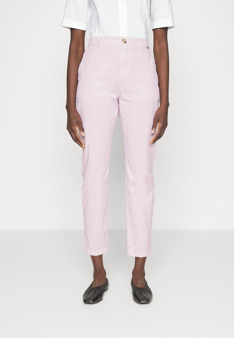 Femme portant un pantalon droit rose clair, un haut blanc à manches courtes et des chaussures plates noires, debout devant un fond uni.