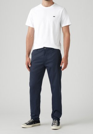 Mann trägt ein weißes kurzärmeliges T-Shirt, marineblaue Hose und schwarze Low-Top-Sneaker mit weißen Schnürsenkeln und steht vor einem einfarbigen Hintergrund.