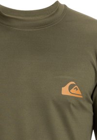 Olivovo zelená dlhá rukávová košeľa z mäkkej a hladkej tkaniny, na ľavej strane hrude s oranžovým logom. Okrúhly výstrih, úzky strih.