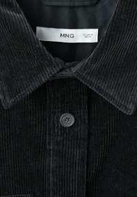 Camicia in corduroy nera con chiusura a bottoni, colletto a punta, strisce testurizzate e un'etichetta che indica marca e taglia.