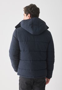 Námornícky modrá puffer bunda s kapucňou na šnúrku, šitím a textúrovanou tkaninou. Má široký strih a predné zapínanie na cvoky.