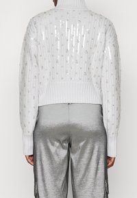 Pull en maille blanc avec motifs pailletés, col montant, poignets et ourlet côtelés. Associé à un pantalon argenté brillant avec poches.