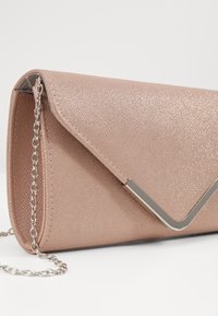 Pochette en or rose scintillante avec rabat triangulaire et détail métallique en forme de V argenté, avec une chaîne argentée.