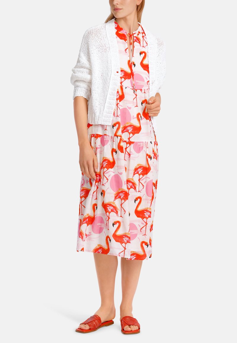 Weißer, offener Strickcardigan über einem bunten Kleid mit einem Flamingomuster in Pink und Rot, ergänzt durch rote flache Sandalen.