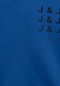 Modrá tkanina s opakujícím se černým textem "J & J" v různých velikostech, s hladkou texturou a rovnoměrnou barvou po celé ploše.