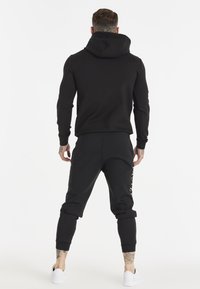 Hombre con tatuajes vistiendo una sudadera con capucha negra y joggers negros, con zapatillas blancas, de pie contra un fondo blanco liso.