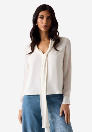 Mujer con cabello largo y oscuro lleva una blusa blanca de manga larga con detalle de corbata en el cuello y pantalones vaqueros azules, de pie frente a un fondo liso.