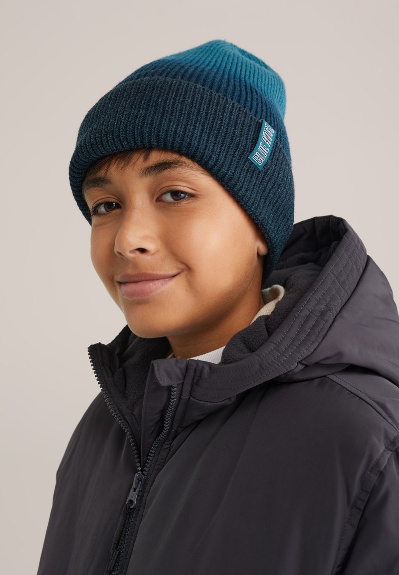 Kötött sapka gradiens kék és teal színben, bordás textúrával és egy kis logópatccsal. Sötét kabáttal viselve.