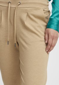 Béžové jogger nohavice vyrobené z textúrovanej látky, s elastickým pásom na šnúrku a bočnými vreckami. Detailný záber na látku a detaily vrecka.