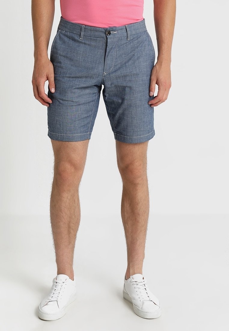 Homme portant un short bleu jusqu'aux genoux, des baskets blanches et une chemise rose, debout devant un fond uni.