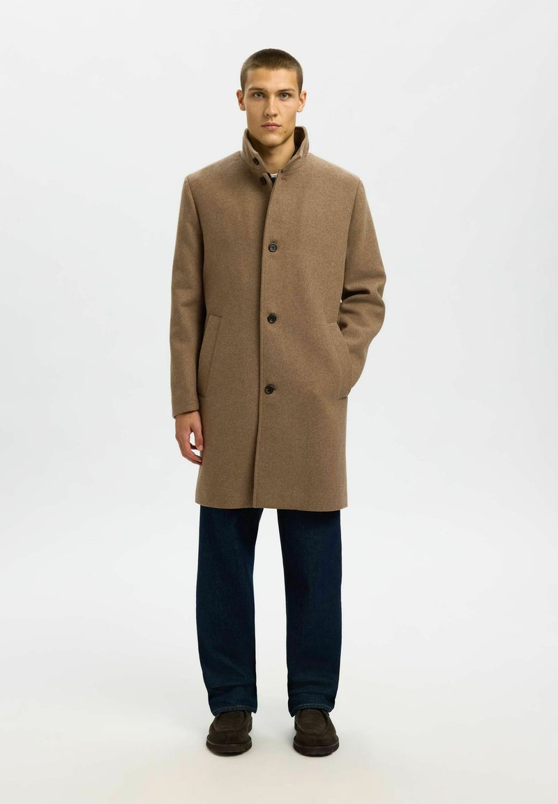 Ung mand iført en brun uldfrakke med knapper, mørke bukser og mørke sko, stående med den ene hånd i lommen mod en hvid baggrund.