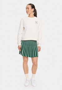 Sweat-shirt blanc avec un graphique vert et des poignets côtelés, associé à une jupe plissée verte avec des rayures verticales blanches. Baskets blanches et chaussettes.