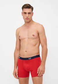 Boxerii roșii din bumbac cu un brâu elastic bleu închis pe care este inscripționat logo-ul "GAP"; țesătura pare netedă și elastică, design ajustat cu o croială de înălțime medie.