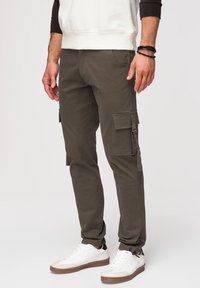 Homme portant un pantalon cargo vert olive, des baskets blanches et un sweat-shirt blanc avec des manches noires, debout avec les mains relâchées.