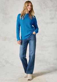Blau gestrickter Pullover mit Rundhalsausschnitt, kombiniert mit hellblauen Schlagjeans. Modell trägt cremefarbene Sneakers vor einem neutralen Hintergrund.