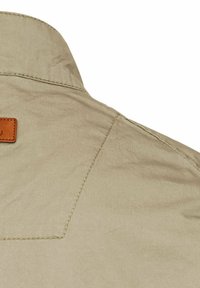 Beige Hemdkragen mit Knopfverschluss und strukturierter Oberfläche, mit einem braunen Lederetikett in der Nähe der Naht und sichtbaren Steppdetails.
