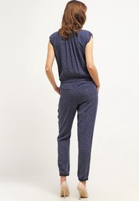Mujer con cabello castaño que lleva una blusa sin mangas de color azul marino con estampado y pantalones a juego, de pie con las manos en los bolsillos, tacones altos beige.