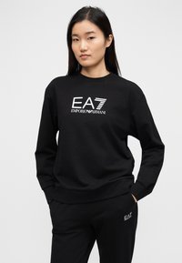 Μαύρο βαμβακερό φούτερ με μακρύ μανίκι και στρογγυλή λαιμόκοψη, με λευκό λογότυπο EA7 Emporio Armani στο μπροστινό μέρος και μαύρο παντελόνι σετ με το ίδιο λογότυπο.