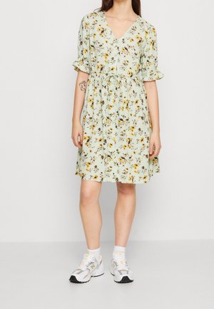 Kvinde iført en lys grøn knælængde kjole med blomsterprint, flæseærmer og hvide sneakers, stående foran en ensfarvet baggrund.