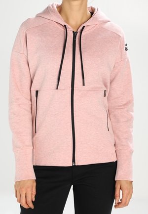 Kobieta ubrana w jasnoróżową bluzę z kapturem na zamek błyskawiczny z czarnymi sznurkami i zamkami, w połączeniu z czarnymi spodniami.