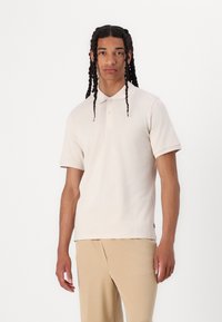 Polo shirt beige chiaro in tessuto liscio, con colletto, due bottoni e maniche corte. Indossato con pantaloni beige chiaro.