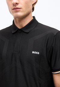 Νέος άνδρας με μαύρο πόλο μπλουζάκι με διακριτικά γεωμετρικά σχέδια και λογότυπο «BOSS» στο στήθος, λευκό φόντο.