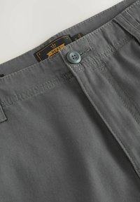 Grau Baumwollhosen-Detail mit geknöpftem Bund und sichtbarem schwarz-gelbem Markenetikett innen am hinteren Bund.