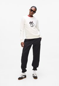 Biała sweatshirt z rysunkowym motywem kota, czarne joggery i czarne okulary przeciwsłoneczne. Ubranie charakteryzuje się teksturowanymi tkaninami i kontrastującymi kolorami.