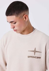 Giovane uomo con un taglio di capelli corto sfumato, che guarda in basso, indossa un maglione a coste beige con un design di stelle e catena sul petto.