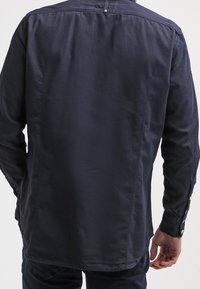 Marineblaues Hemd mit einem geknöpften Kragen, langen Ärmeln und Seitennähten. Es verfügt über eine glatte Baumwollstruktur und eine einzelne Knopfleiste am Nacken hinten.