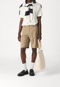 Beige Shorts, weißes T-Shirt, marineblau- und cremefarben gestreifter Pullover über die Schultern gelegt. Schwarze Schuhe, weiße Socken, hält eine hellbraune Tragetasche.