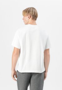 Biela bavlnená tričko, krátke rukávy, klasický výstrih, voľný strih, s logom na zadnej strane. Spárované so šedými textúrovanými teplákmi.
