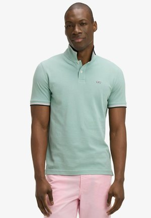 Homme portant un polo à manches courtes vert menthe avec col relevé et un short rose, debout devant un fond blanc uni.