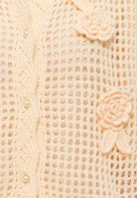 Πλεκτή ζακέτα σε ανοιχτό ροδακινί χρώμα με σχέδιο από δίχτυ, διακοσμημένη με εφαπτόμενα λουλουδάτα μοτίβα και κουμπιά από μαργαριτάρια κατά μήκος του μπροστινού μέρους.