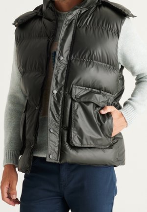 Man die een grijze doorgestikte mouwloze bodywarmer draagt over een lichtgrijze trui, met zijn hand in een groot voorvak, gecombineerd met donkerblauwe broek.