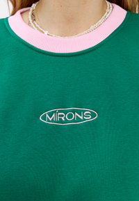 Felpa verde con colletto a coste rosa. Presenta la parola ricamata "MIRONS" di un rosa chiaro sul davanti. Tessuto morbido.