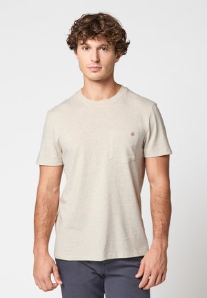 Jeune homme aux cheveux bouclés portant un t-shirt beige clair à manches courtes avec un petit logo de palmier sur la poche poitrine.