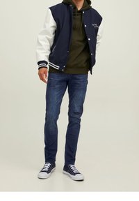 Marinblå och vit varsityjacka med tryckknappar, i kombination med en grön huvtröja, slitna blå jeans och marinblå sneakers med vita sulor.