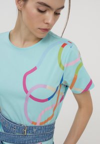 T-shirt azzurro chiaro con motivi astratti colorati a linee nei toni di rosa, arancione e verde. Tessuto morbido, maniche corte, scollatura rotonda.