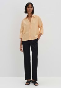 Camicia a maniche lunghe arancione chiaro, con bottone e tessuto testurizzato, abbinata a pantaloni neri a gamba dritta.