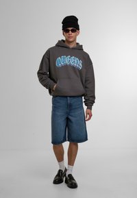 Grå hettegenser med "QUEENS" grafikk, blå skrift; denimshorts; svarte sko; hvite sokker; svart lue; står mot en grå bakgrunn.