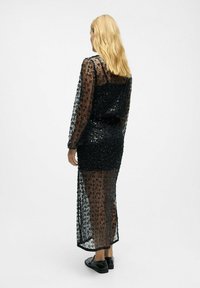 Schwarzes, mit Pailletten besetztes Kleid mit langen, durchsichtigen Ärmeln, tailliertem Oberteil und langem Rock. Der Stoff zeigt ein florales Muster mit glänzender Oberfläche.