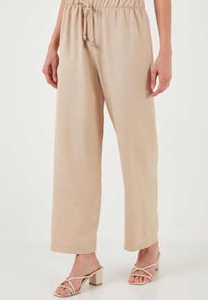 Beige Leinenhose mit einem Kordelzugbund, weiter Beinform und strukturierter Optik, kombiniert mit beigen Sandalen mit Blockabsatz und Riemchen.