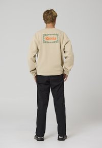 Beiger Pullover mit grünem und orangefarbenem "Rivvia"-Schriftzug und floralem Design auf der Rückseite, kombiniert mit schwarzer Hose und schwarzen Schuhen.